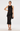 A model wearing a sleeveless black midi dress with ruffled trim and a tie-neck detail, holding a small black clutch against a plain white studio background.