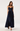 A woman modeling a navy blue maxi dress with a pleated skirt, lace-trimmed spaghetti straps, and a sweetheart neckline, photographed against a plain light gray studio background.