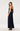 A woman modeling a navy blue pleated maxi dress with thin straps, a low back cutout, and large bow details on the back, shown against a plain light gray studio background.