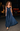 A woman wearing a floor-length navy blue pleated gown with a lace-trimmed bodice and thin straps, walking outdoors at night on a city street.