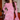 A woman wearing a pink lace long-sleeve mini dress with flared cuffs, standing in a restaurant or bar setting with wooden shelves and bottles in the background.
