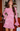 A woman wearing a pink lace long-sleeve mini dress with flared cuffs, standing in a restaurant or bar setting with wooden shelves and bottles in the background.