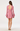 A woman shown from the back wearing a pink textured long-sleeve mini dress with a large open back, flared cuffs, and a ruffled hem, paired with white heeled sandals against a plain light background.