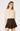 A woman modeling a cream long-sleeve blouse with a lace ruffled collar and bow tie detail, paired with a dark brown pleated mini skirt, against a plain light gray studio background.