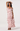 A woman modeling a light pink ruffled maxi dress with a dusty rose floral print, photographed against a plain light gray studio background.