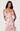 A woman modeling a light pink ruffled spaghetti-strap dress with a dusty rose floral print against a plain light gray studio background.