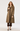 A woman modeling a mid-length brown and beige houndstooth pattern trench coat over a black top, dark brown cropped pants, and black ankle-strap heels, photographed against a light gray studio background.