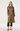A woman modeling a mid-length brown and beige checkered trench coat with a matching waist belt, paired with dark brown cropped pants and black heels, against a plain light gray studio background.