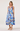 A woman modeling a sleeveless midi dress with a blue and purple floral print on a white background, paired with beige strappy high-heeled sandals, against a light gray studio backdrop.