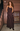A woman with curly brown hair models a dark brown ruffled maxi dress with spaghetti straps, standing on a city sidewalk in front of a brown brick building.
