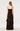 A woman viewed from the back wearing a dark brown spaghetti-strap maxi dress with ruffle details and an asymmetrical layered hem, photographed against a plain light gray studio background.