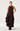 A woman modeling a sleeveless dark brown midi dress with a high neck, front tie detail, and a tiered ruffled skirt, paired with dark brown pointed boots against a light gray studio background.
