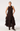 A woman modeling a sleeveless dark brown midi dress with a high neck, front tie detail, and a tiered ruffled skirt, paired with dark brown heeled ankle boots against a plain light gray studio background.