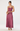 A woman modeling a sleeveless maxi dress with a structured, corset-style bodice and a flowing satin-like skirt in a dusty rose or mauve shade, paired with nude high heels and a small gold clutch, against a plain light gray studio background.