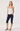 A woman modeling a white halter top with a small black polka dot pattern, paired with navy blue capri pants and black slide sandals, against a light gray studio background.