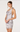 A woman modeling a light silver satin halter mini dress with a draped neckline, lace trim at the hem, and a tie detail at the shoulder, set against a plain light gray studio background.