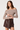 A woman modeling an off-the-shoulder long-sleeve top with a brown and beige geometric pattern, paired with a dark brown faux-leather wrap skirt, against a plain light background.