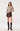 A model wearing a taupe knit off-the-shoulder sweater, a dark brown faux-leather wrap mini skirt, and black knee-high leather boots, photographed against a seamless white studio background.