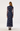 A woman viewed from the back wearing a navy midi dress with sheer long sleeves and a blue floral pattern, paired with black high-heeled shoes against a plain white studio background.