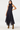 A woman modeling a sleeveless high-neck midi dress with a navy blue base and small white polka dots, paired with brown suede ankle boots against a plain light gray studio background.