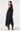 A woman modeling a sleeveless navy blue high-low dress with a small white polka dot pattern, shown from a three-quarters back view against a light gray studio background.