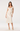 A model wearing a cream-colored two-piece dress set consisting of a ruffled short-sleeve crop top with a front bow and a matching midi skirt with a subtle jacquard pattern, paired with cream high heels against a plain white studio background.