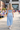 A woman viewed from behind walking outdoors on a sunny day, wearing a light blue matching two-piece outfit consisting of a cropped spaghetti-strap top and a high-waisted, wide-leg skirt with a thin red stripe detail at the waist.