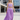 A woman modeling a lavender two-piece outfit consisting of a cropped tank top and matching high-waisted maxi skirt, set against a modern concrete and glass stadium background.