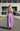 A woman viewed from behind wearing a lavender purple two-piece set consisting of a cropped camisole top and a tiered maxi skirt, walking on an outdoor paved path in front of a modern concrete building.