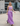 A woman modeling a lavender two-piece set consisting of a cropped tank top and a matching maxi skirt, walking outdoors in front of a modern stadium and convention center.
