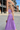 A woman wearing a lavender purple two-piece set with white geometric embroidery, walking outdoors in front of a brick building.