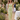 Woman in a green checkered dress standing in a garden setting with floral decorations and people in the background.