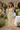 Woman in a green checkered dress standing in a garden setting with floral decorations and people in the background.