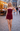 A woman stands on a cobblestone street wearing a strapless burgundy mini dress with a ruched bodice and asymmetrical hem. The background shows a softly blurred outdoor cafe and city street at dusk.