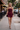 A blonde woman walking on a city sidewalk wearing a strapless, mini-length burgundy dress with a draped, textured bodice and flared skirt. She is holding a small black clutch and wearing black strappy high-heeled sandals.