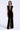 A woman modeling a sleeveless black velvet floor-length gown with a plunging V-neckline, sheer mesh panel at the neckline, and a high front slit, photographed against a light gray studio background.