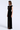 A woman modeling a floor-length black gown with a sheer mesh insert at the neckline and a high front slit, photographed against a clean light gray studio background.