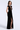 A woman modeling a floor-length black evening gown with a plunging V-neckline, sheer mesh panels, and a high thigh slit, photographed against a clean light gray studio background.