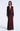 A woman modeling a floor-length, long-sleeve evening gown in deep burgundy with a sheer mesh overlay and gathered ruching at the waist, photographed against a clean light gray studio background.