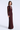 A woman modeling a floor-length, deep V-neck evening gown in a rich burgundy color with sheer long sleeves and a textured, shimmering fabric, set against a light gray studio background.