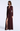 A woman modeling a floor-length burgundy gown with sheer long sleeves, a deep V-neckline, and a high thigh slit, photographed against a clean light gray studio background.