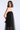 A woman modeling a strapless black tulle dress with a wide dark brown or very deep black satin-like band across the bust, photographed against a clean light gray studio background.
