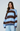 A woman wearing a loose, oversized knit sweater with wide horizontal stripes in light blue, dark brown, and navy blue, paired with light blue pants, against a plain light gray studio background.