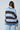 A woman shown from behind wearing a loose knit sweater with wide horizontal stripes in light blue, dark brown, and navy blue, paired with light blue pants against a plain light gray studio background.