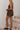 A woman modeling a brown strapless tube top and matching shorts set, standing on rustic brick stairs against a white wall.