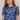 A woman with long red hair wearing a short-sleeve blue mesh top with a white daisy floral pattern, photographed against a plain light beige studio background.