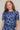 A woman with long red hair wearing a short-sleeve blue mesh top with a white daisy floral pattern, photographed against a plain light beige studio background.