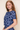 A woman with long light brown hair wearing a blue and white floral lace top, photographed against a plain beige studio background.