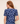 A woman shown from the back wearing a navy blue short-sleeve top with a light blue floral lace pattern and a small keyhole cutout at the neckline, against a plain beige background.