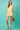 Woman wearing a light green outfit holding a hat against a teal background